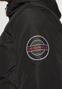 Giacca nera con un patch rotondo sulla manica superiore che riporta "JACK&JONES," "ORIGINALS," e "SINCE 1989" in rosso e blu.