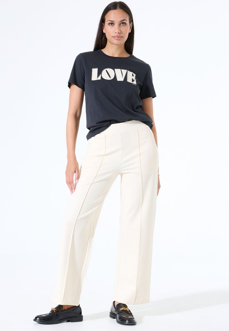 Camiseta negra de manga corta con un gráfico de "AMOR", combinada con pantalones de pierna ancha color crema. El modelo lleva zapatos tipo mocasín negros con detalle de cadena dorada.