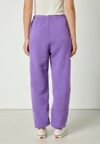 Violet spalvos pūkuotos sportinės kelnės su laisvu siluetu, turi elastinį juosmenį ir subtilią tekstūrą, derinamos su baltomis sportinėmis bateliais.