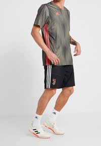 Uomo che indossa una maglia da calcio Adidas grigia e rossa, pantaloni corti neri della Juventus, calze bianche Adidas e scarpe bianche con strisce nere.