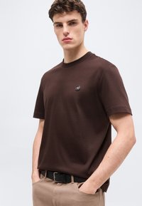 Νεαρός άνδρας που φοράει κοντομάνικη καφέ σκουρόχρωμη μπλούζα με μικρό στρογγυλό λογότυπο στο στήθος, μπεζ παντελόνι και μαύρη ζώνη, με τα χέρια στις τσέπες.