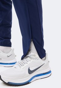 Μπλε σκούρο παντελόνι με φερμουάρ στον αστράγαλο μισά ανοιχτό πάνω από λευκά αθλητικά Nike Air Zoom με μπλε σόλα και λευκές κάλτσες.