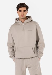 Beige hoodie med lynlås, som har en frontlomme, hætte med snore og en blød, glat tekstur. Minimalistisk branding synlig på brystet.