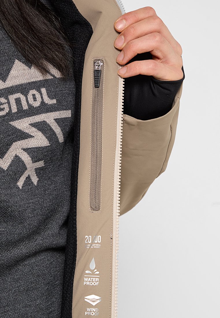 Beige Jacke mit einer Reißverschlusstasche, mit wasser- und winddichter Kennzeichnung. Innen sieht man einen dunkelgrauen Pullover mit einem Logo und Design.