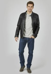 Zwarte leren jas met ritszakken, lichtgrijze henley-shirt, blauwe spijkerbroek en bruine veterschoenen. Model staat casual.