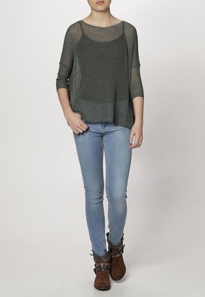 Szary, luźny sweter z szerokim dekoltem i rękawami o długości trzy czwarte, połączony z jasnoniebieskimi dżinsami typu skinny i brązowymi botkami za kostkę.