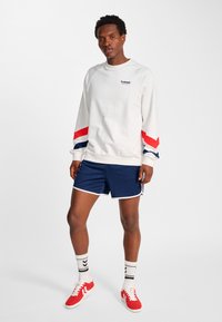Vit långärmad sweatshirt med röda och blå ränder, kombinerad med marinblå shorts med vit kant, röda sneakers och mönstrade strumpor.