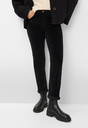 Eine Person trägt schwarze Cord-Hosen mit umgeschlagenen Säumen, schwarze klobige Lederstiefel, ein cremfarbenes Oberteil und eine schwarze Jacke und steht an einer weißen Wand.