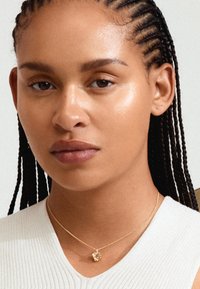 Goldene Halskette mit einem kleinen Würfel-Anhänger, getragen von einer Person mit geflochtenem Haar, vor einem neutralen Hintergrund. Einfaches Design und glatte Textur.