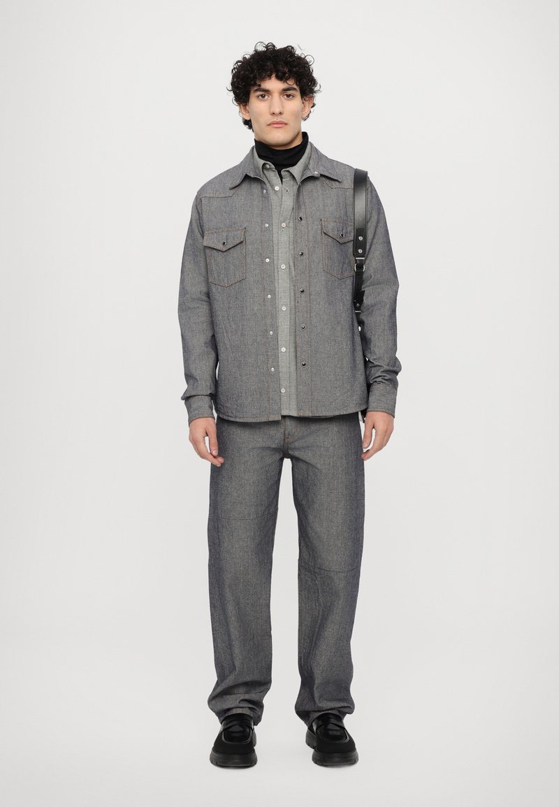 Grå denimjacka med två bröstfickor, knäppning med knappar och kontrasterande sömmar, kombinerad med matchande grå byxor och svarta skor.