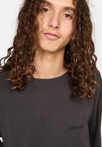 Dunkelgraues Langarmshirt aus weichem Stoff, mit Rundhalsausschnitt und kleinem schwarzen Logo auf der Brust. Lockige Haare sind teilweise sichtbar.