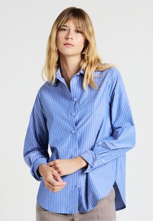 Giovane donna che indossa una camicia a righe verticali blu e bianche, pantaloni beige, con le mani unite davanti, sfondo neutro.