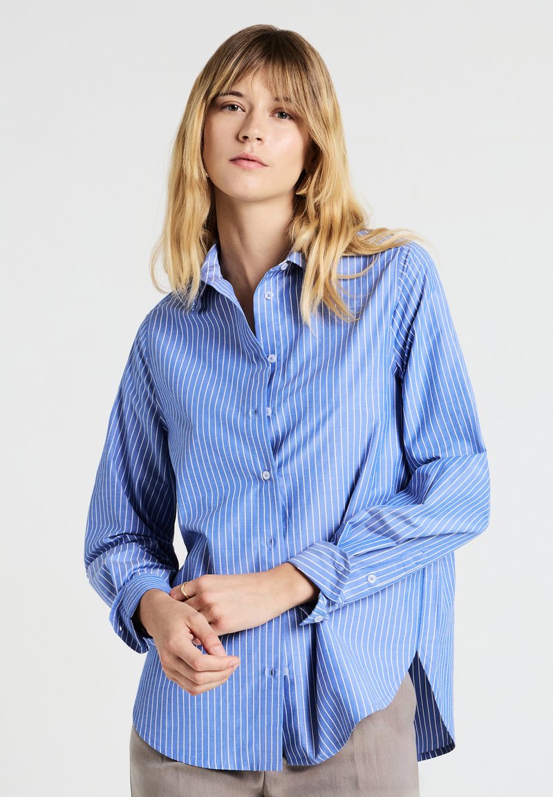 Giovane donna che indossa una camicia a righe verticali blu e bianche, pantaloni beige, con le mani unite davanti, sfondo neutro.