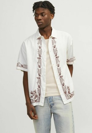 Chemise blanche à manches courtes avec broderie florale brune sur le devant et les manches. Elle présente un design à boutons et un tissu léger.