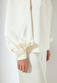 Cream-colored blouse with voluminous sleeves and buttoned cuffs. Features a smooth, shiny fabric and a relaxed fit.