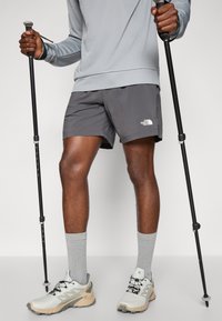 Harmaa pitkähihainen paita, harmaat shortsit, joissa valkoinen logo, harmaat sukat ja beiget vaelluskengät. Molemmissa käsissä vaelluskeppi.