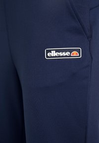 Marina atletiska byxor tillverkade av texturerat tyg, med en broderad ellesse-logotyp i vitt och orange på vänster sida.