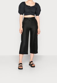 Top negro corto con mangas abullonadas, combinado con pantalones negros de pierna ancha y cintura alta. La modelo lleva sandalias de plataforma negras. Texturas suaves.