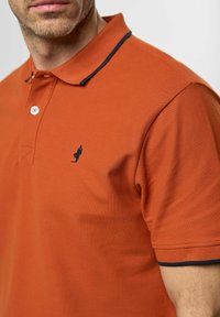 Narančasta polo majica s teksturiranim završetkom, s tamnoplavim ovratnikom i malim tamnoplavim logom na lijevoj strani grudi. Zatvaranje gumbom na ovratniku.