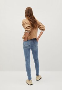 Suéter beige de canalé con mangas abullonadas, jeans ajustados azules y zapatillas de color claro. Bolsillos traseros visibles en los jeans, textura suave.