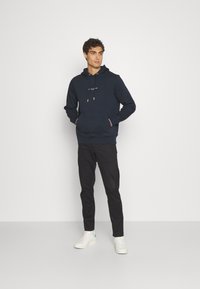 Felpa navy con tasca frontale, cappuccio con cordino e testo bianco sul petto. Abbinata a pantaloni neri e sneakers bianche.