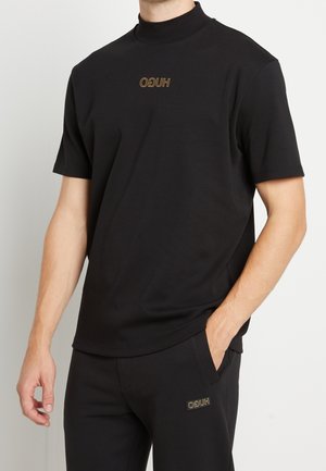 Homme portant une chemise noire à manches courtes et un pantalon avec de petits logos dorés "HUGO" sur la poitrine et la partie supérieure de la cuisse, main dans la poche.