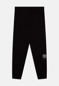 Pantalon de jogging noir avec une taille élastique, poches latérales, texture lisse et un écusson en métal argenté sur une jambe.