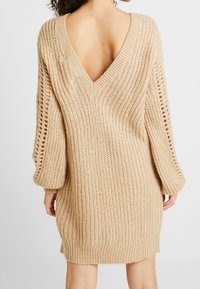 Abito in maglia beige con scollatura a V profonda sul retro, texture a coste, maniche sfuse e tagli laterali. Tessuto morbido, lunghezza fino a metà coscia.