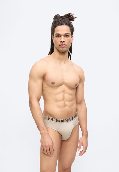 Μυώδης άντρας με μακριές κοτσίδες, φορώντας μπεζ εσώρουχα Emporio Armani, στέκεται μπροστά σε απλό ανοιχτό γκρι φόντο, κοιτάζοντας προς τα εμπρός.