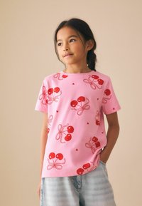 Camiseta de manga corta rosa hecha de algodón. Presenta gráficos de cerezas rojas con lazos, creando un patrón divertido. Diseño suave y casual.