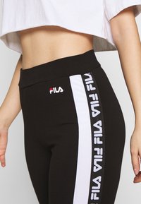 Černé legíny s vysokým pasem, zdobené bílými postranními pruhy a malým logem FILA v červené a bílé barvě. Měkká, elastická tkanina.