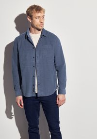 Men's blue button-up shirt in soft fabric, featuring a chest pocket, black buttons, and long sleeves. Paired with dark jeans.