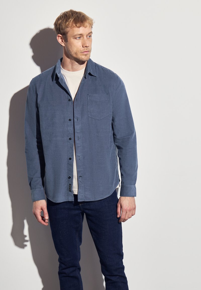 Men's blue button-up shirt in soft fabric, featuring a chest pocket, black buttons, and long sleeves. Paired with dark jeans.