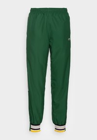 Pantalones deportivos verdes de tela suave, con cinturilla elástica, puños a rayas en negro, blanco y amarillo, y un detalle de logo en el lateral.