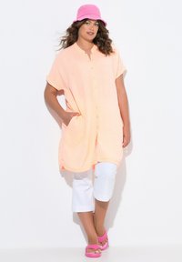 Tunic color pesca con bottoni e maniche corte, abbinata a pantaloni bianchi cropped e sandali rosa, completata da un cappello a secchiello rosa.