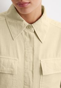 Light beige button-up shirt made of textured fabric, featuring a pointed collar, dual chest pockets, and visible stitching details.