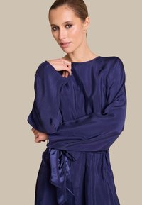 Langermet kjole i dyp marineblå sateng med en avslappet passform, som har et bånd i midjen og rund hals. Jevn tekstur.