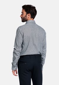 Man die met zijn rug naar de camera staat, draagt een lichtgrijze geblokte blouse met lange mouwen en donkere marineblauwe broeken tegen een effen witte achtergrond.