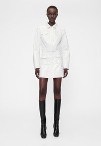 Bela kratka obleka iz lakiranega usnja z ovratnikom, sprednjim zapenjanjem na gumbe, dvema žepoma na prsih in dolgimi rokavi. Oblečena s črnimi škornji do kolen.