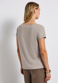 Camiseta corta de color beige claro con un ajuste relajado, cuello redondo y mangas ligeramente enrolladas, combinada con pantalones marrones y un accesorio dorado.