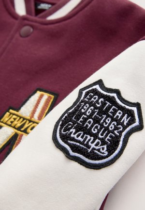 Vinrød varsityjakke med cremefarvede ærmer. Har broderede mærker, herunder en 'NEW YORK'-logo og et skjold med teksten "EASTERN 1961-1962 CHAMPS."