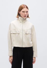 Κοντ ό κρεμ σακάκι από βαμβακερό μείγμα, με ψηλό γιακά, δύο τσέπες στο στήθος και φερμουάρ μπροστά, με ομαλή υφή.