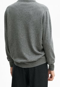 Person, die einen grauen Langarmpullover und schwarze Hose trägt, von hinten vor einem weißen Hintergrund gesehen.