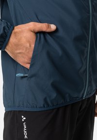Leichte blaue Jacke mit Reißverschlusstasche und strukturiertem Stoff. Verfügt über einen Kordelzug und wird mit schwarzen Hosen kombiniert, die ein Logo zeigen.