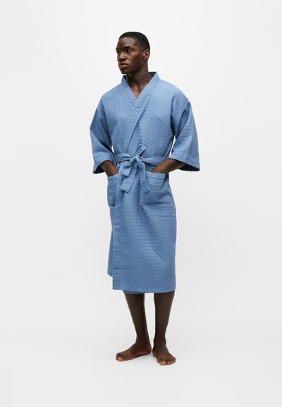 Άνδρας που στέκεται ξυπόλητος, φοράει μπλε μπαντό με υφή βάφλας μέχρι το γόνατο, με μπροστινές τσέπες και δεμένη ζώνη, κοιτάζοντας προς τα πλάγια.