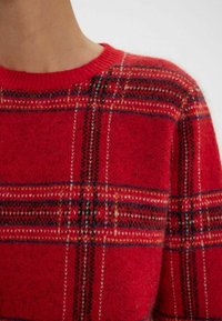 Pull en tartan rouge à col rond. Il présente un mélange de laine doux et texturé, avec des lignes noires et blanches contrastantes formant un motif à carreaux.
