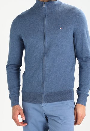 Man wearing a blue zip-up sweater with a small logo on the chest and light blue pants, standing with hands relaxed by sides.