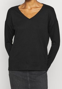 Haut noir à manches longues avec encolure en V, fabriqué en tissu doux, avec épaules tombantes et coupe décontractée. L'ourlet est droit et légèrement plus long à l'arrière.