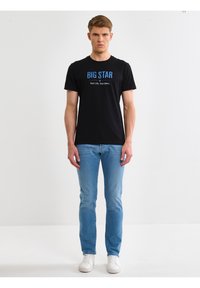 Mladić stoji uspravno, nosi crnu majicu s natpisom "BIG STAR", svijetloplave traperice i bijele tenisice, na jednostavnoj bijeloj pozadini.