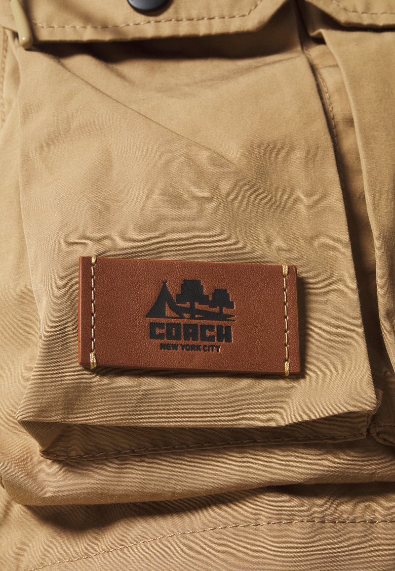 Un tissu beige présente une étiquette en cuir avec "COACH NEW YORK CITY" en noir, entourée d'une bordure cousue. L'étiquette comprend un design graphique.